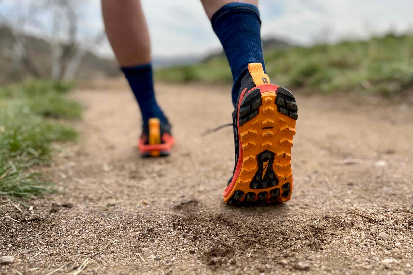Close up from behind of a person wearing trail running sheos walking down a trail. the lug pattern is visible on one shoe.