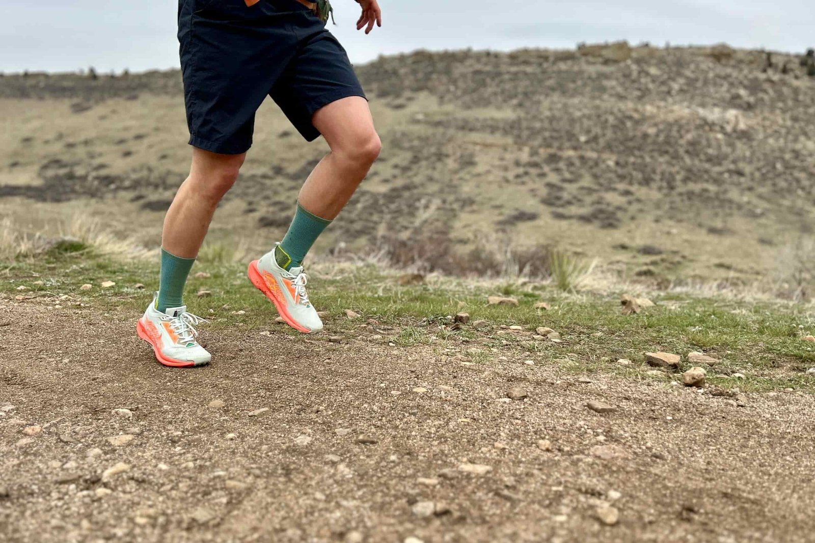 A person from the waist down runs on a dirt road in a mountain setting.