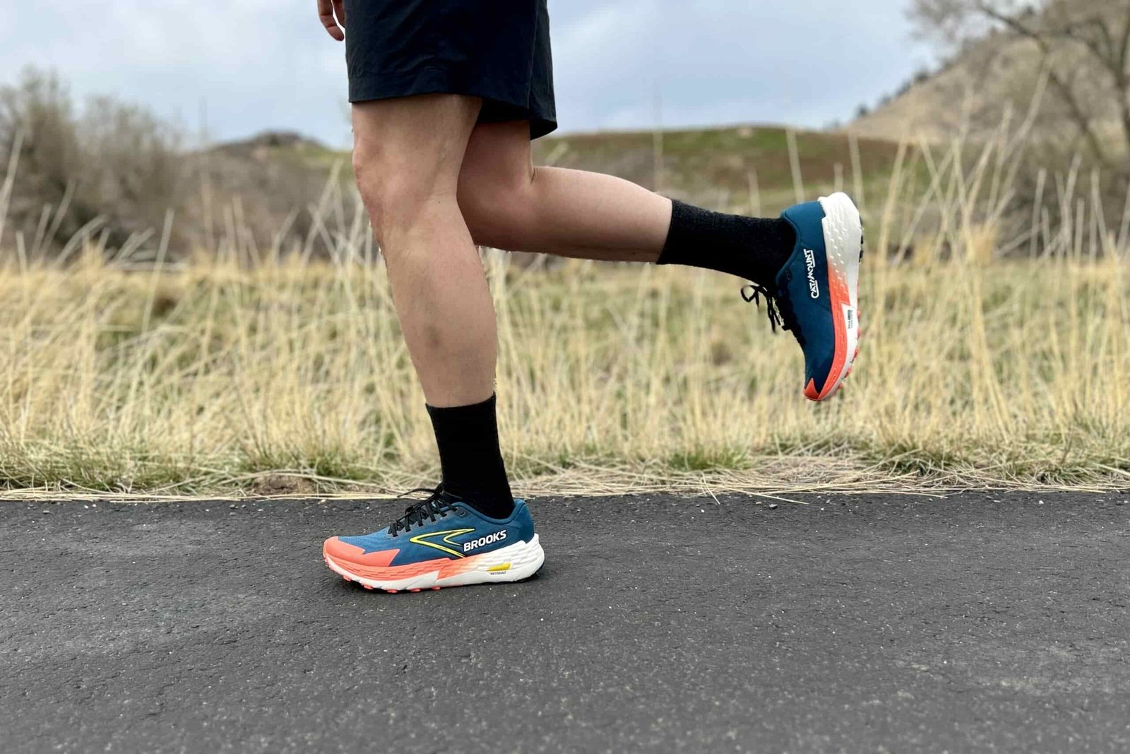 Close up image from the waist down of a person in trail running shoes and shorts running on a road with tall grass at the edge.