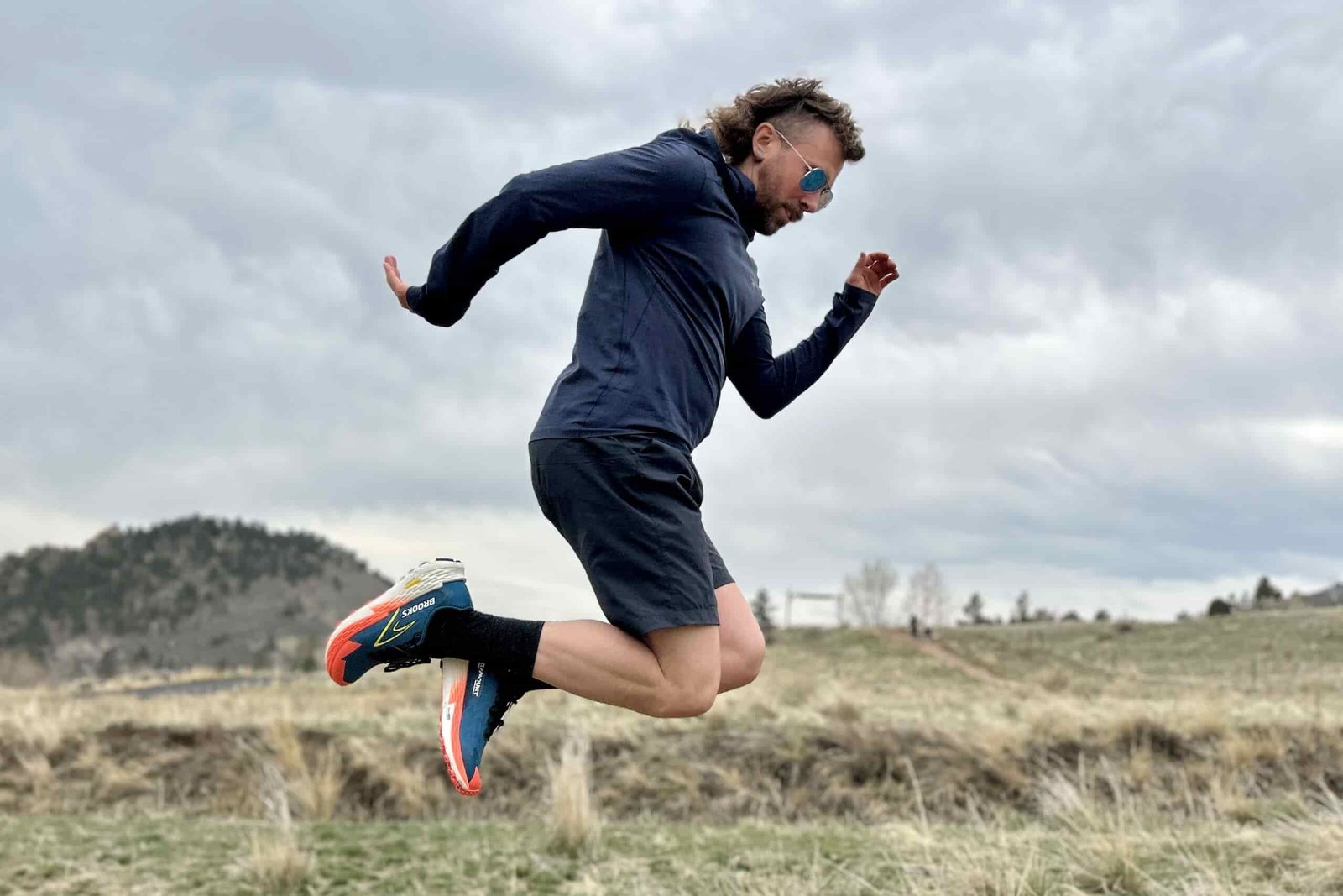 A man jumping in the air wearing trail running attire under a moody, cloudy sky.