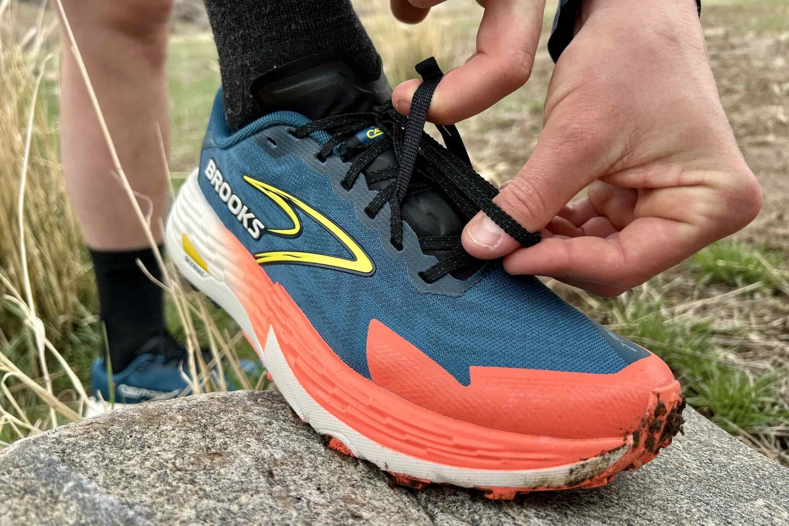 Close up image of a person tucking the shoelaces of a trail running shoe under an elastic strap on the lacing saddle.