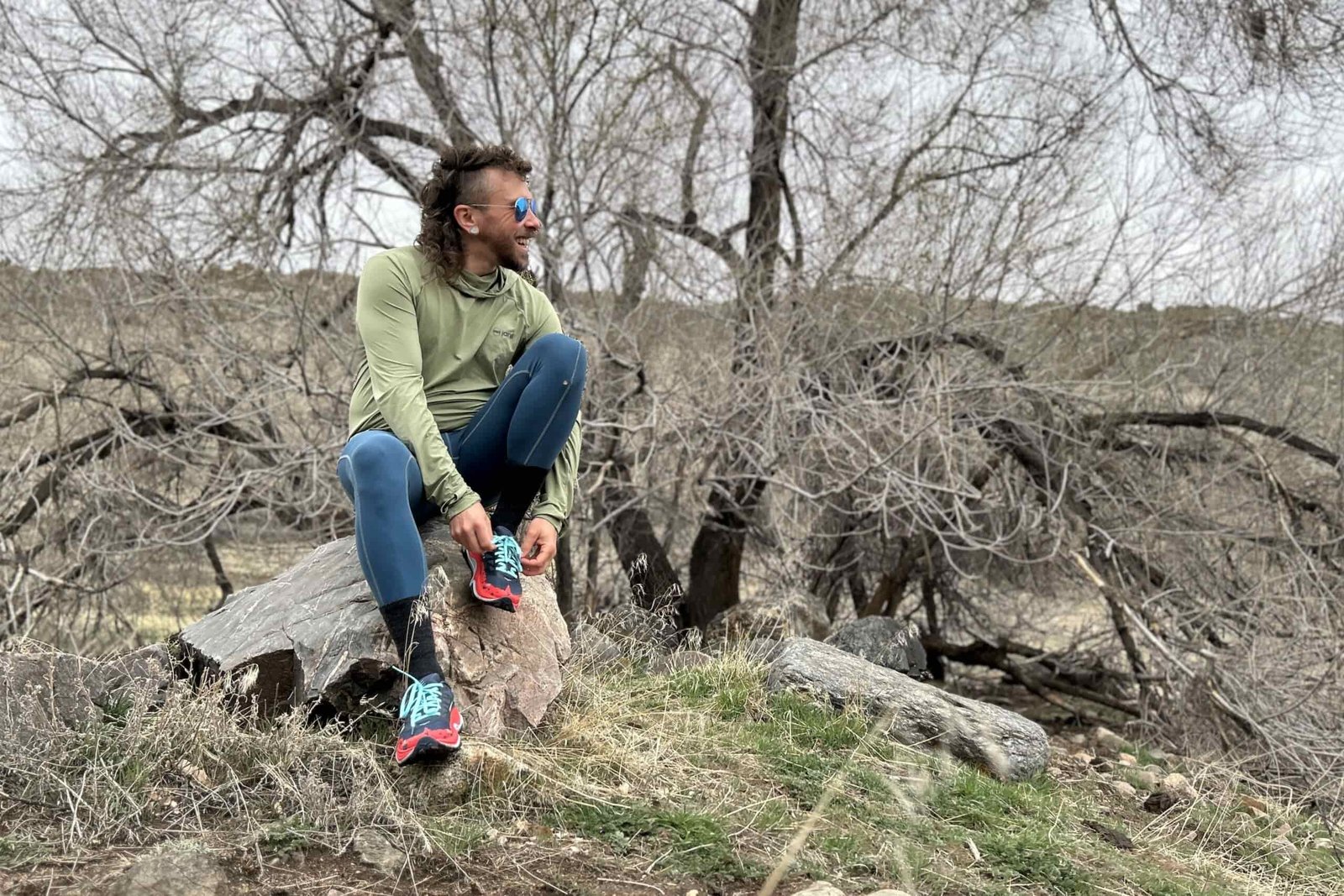 A man sits on a rock in a wooded landscape tying his shoes and looking off in the distance.
