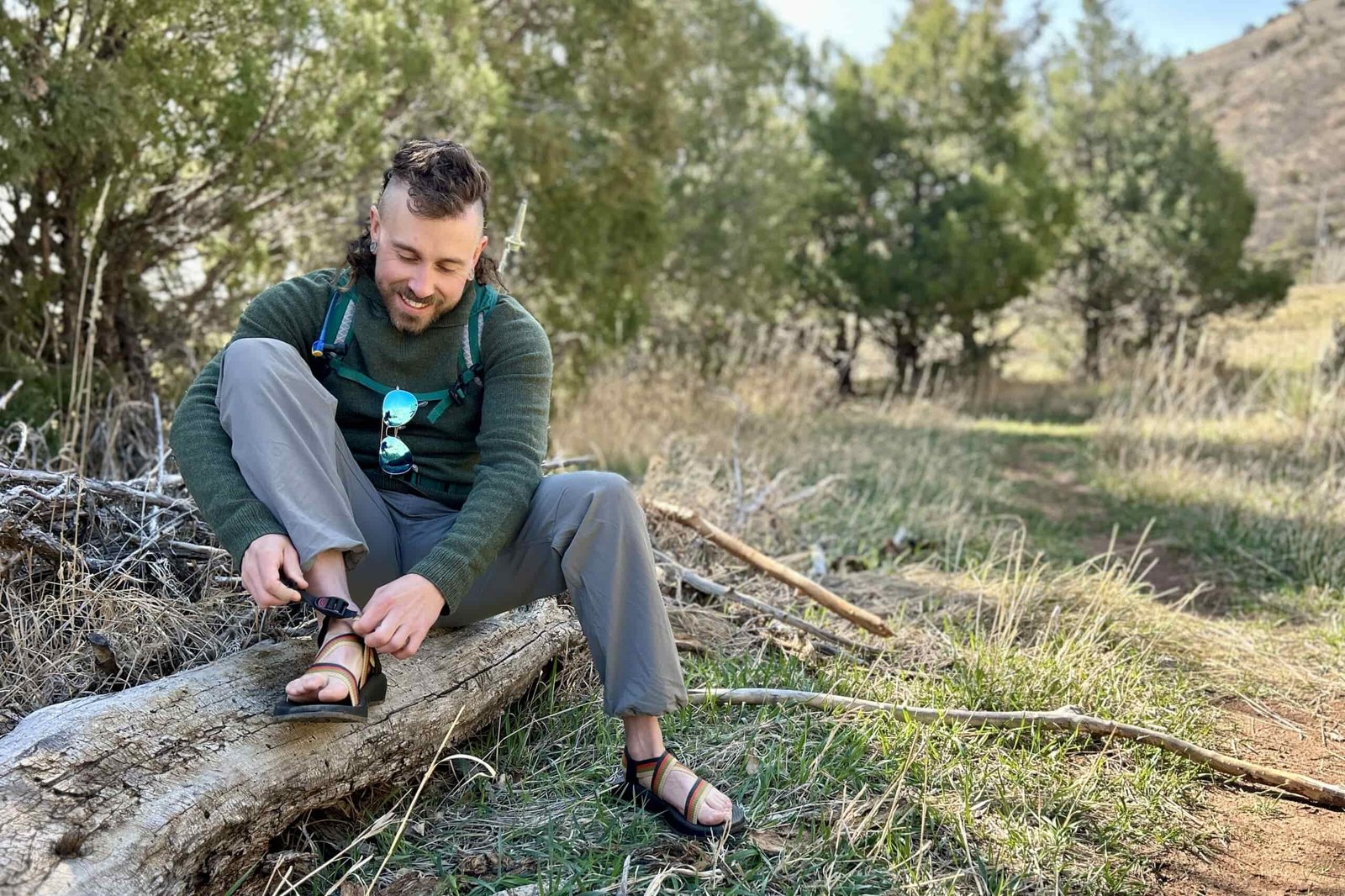 A man sits on a log, smiling as he adjusts a pair of sandals