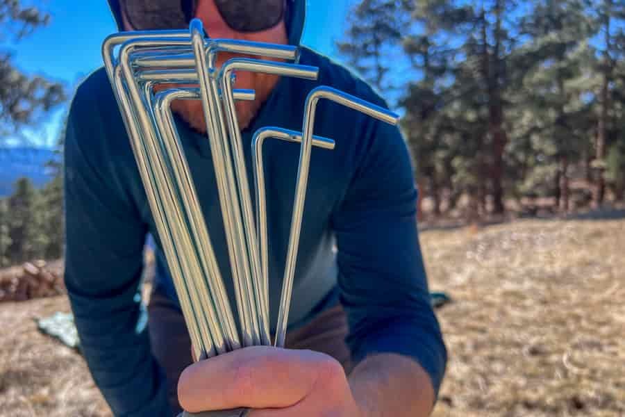 A man holds silver tent stakes up the the camera. The man and the trees in the background are blurry but the tent stakes are in focus.