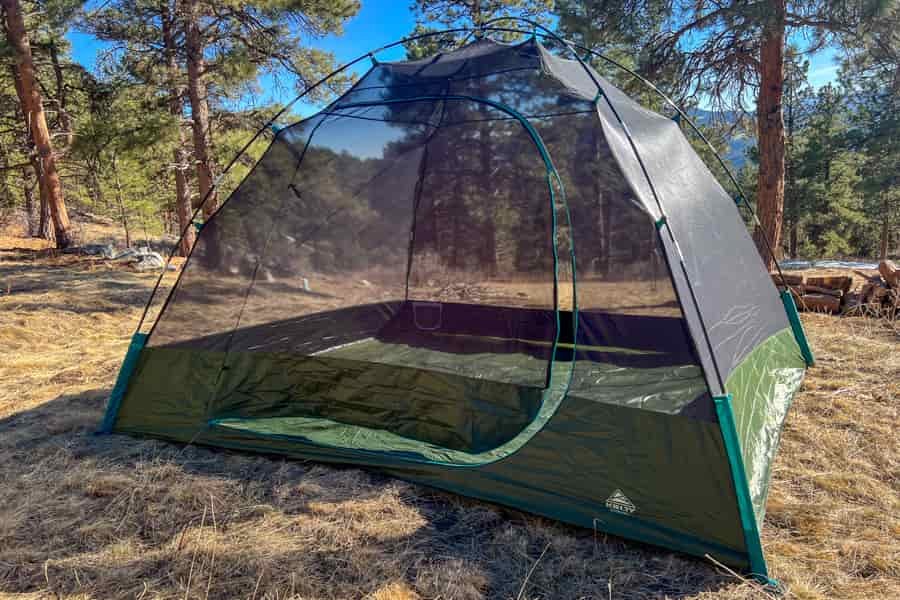 A mesh tent sits without it's rainfly in e field of brown grass. In the background there are pine trees and blue sky.