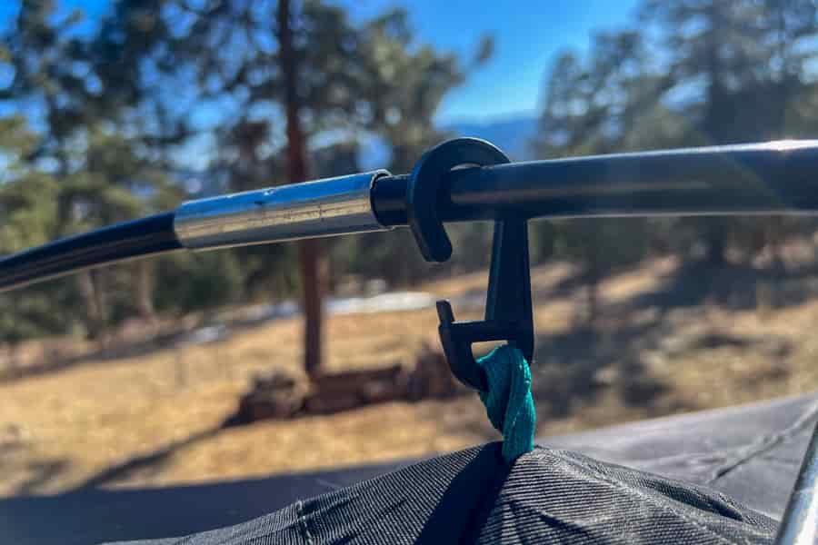a plastic clip is clipped to a tent pole. The background is blurry and consists of pine trees ans blue sky.
