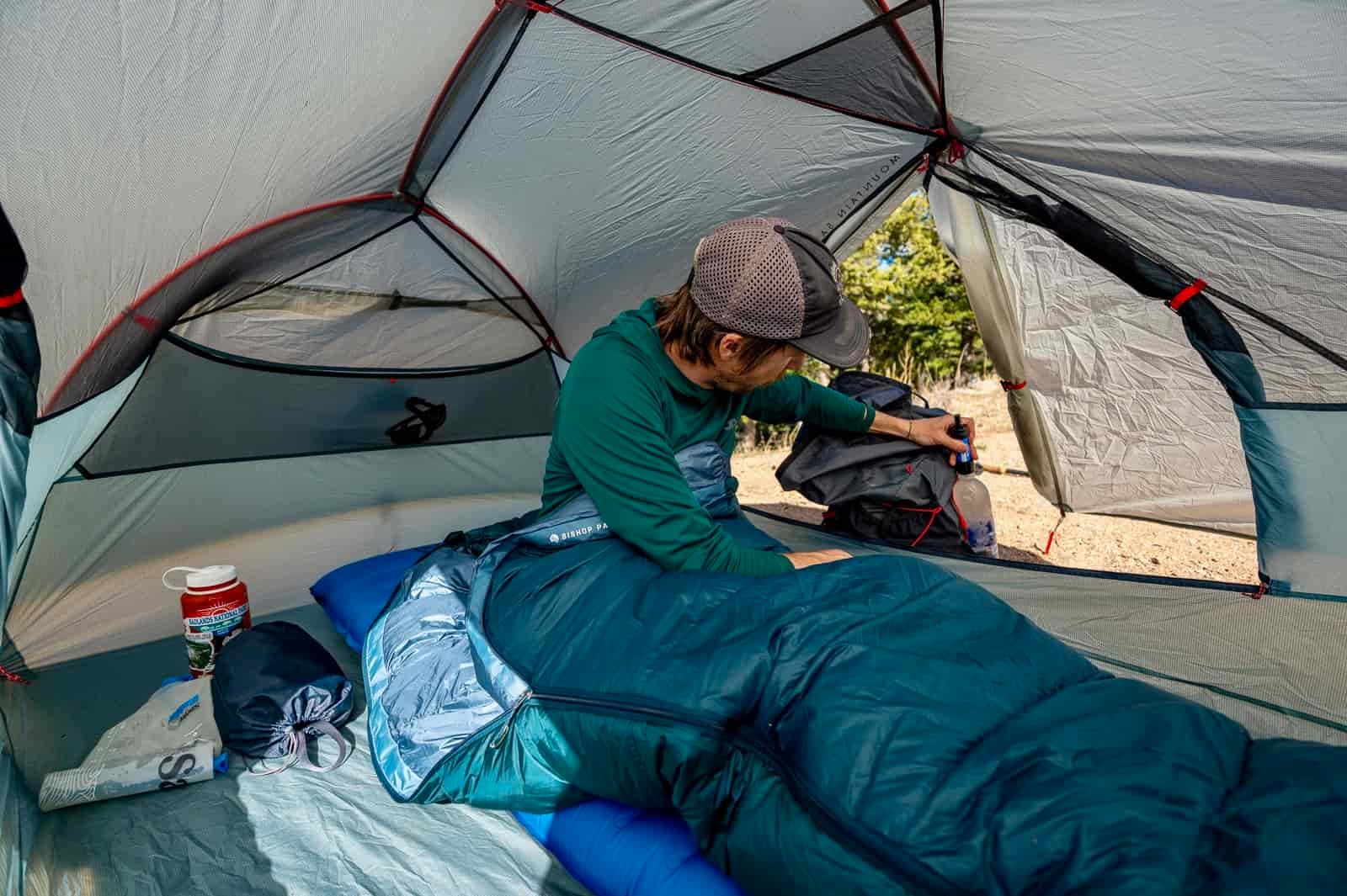 a hiker sits in a tent in a sleeping bag while he reaches for his water bottle.