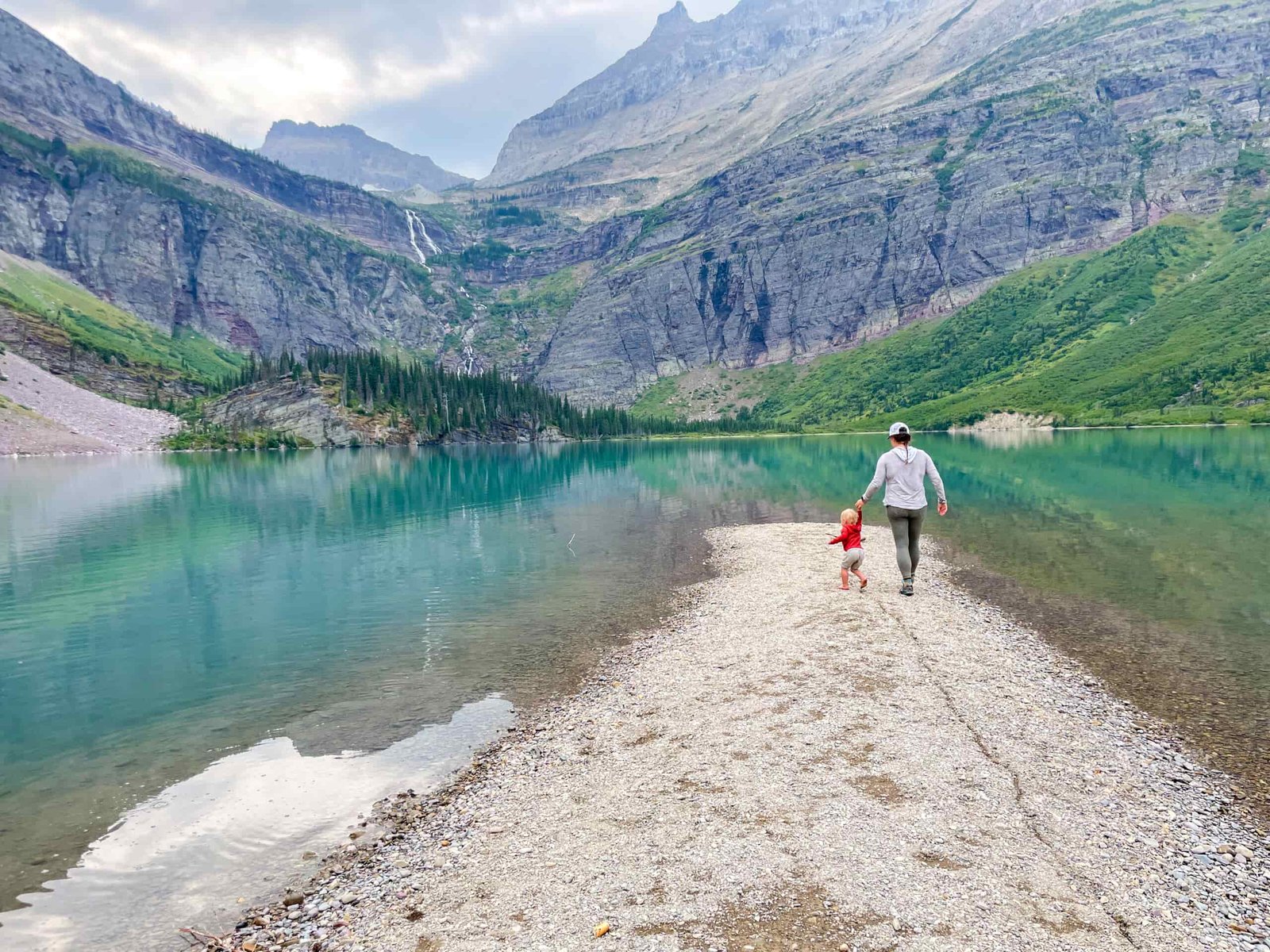 a mom and kid walk along a sandbar in a lake