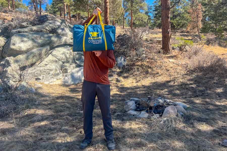 A man holding a tent bag in front of his face. There are pine trees in the background.