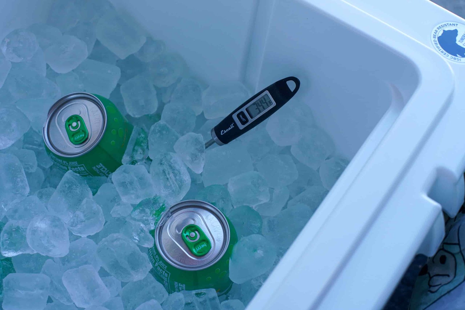 A thermometer reading 34.4 degrees fahrenheit sticks out of a bed of ice in the corner of the YETI Tundra 65 cooler. Two canned drinks poke out of the ice.