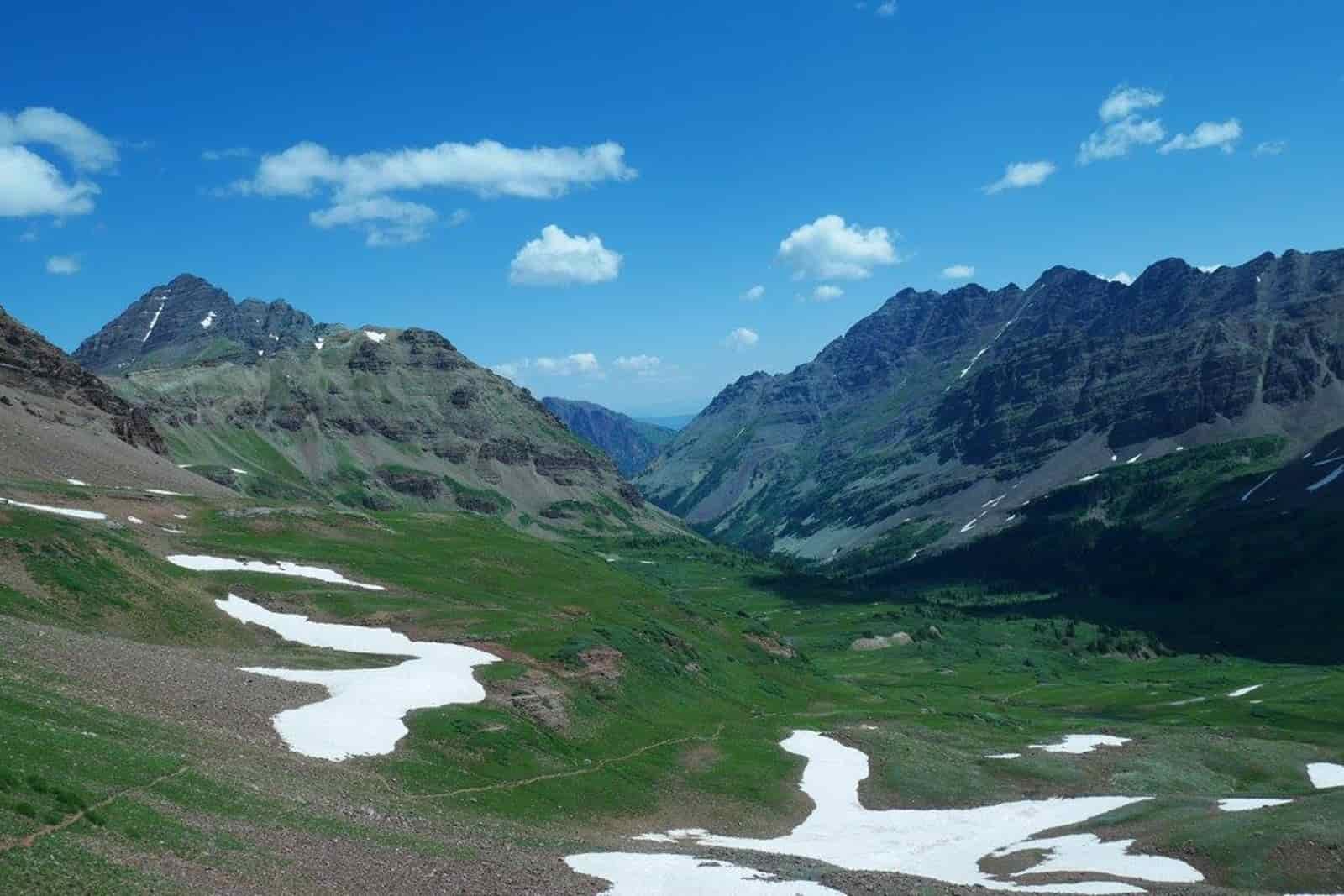 View of some ragged mountains with a trail running down into the valley. It is mostly open due to being above tree line with some patches of snow.