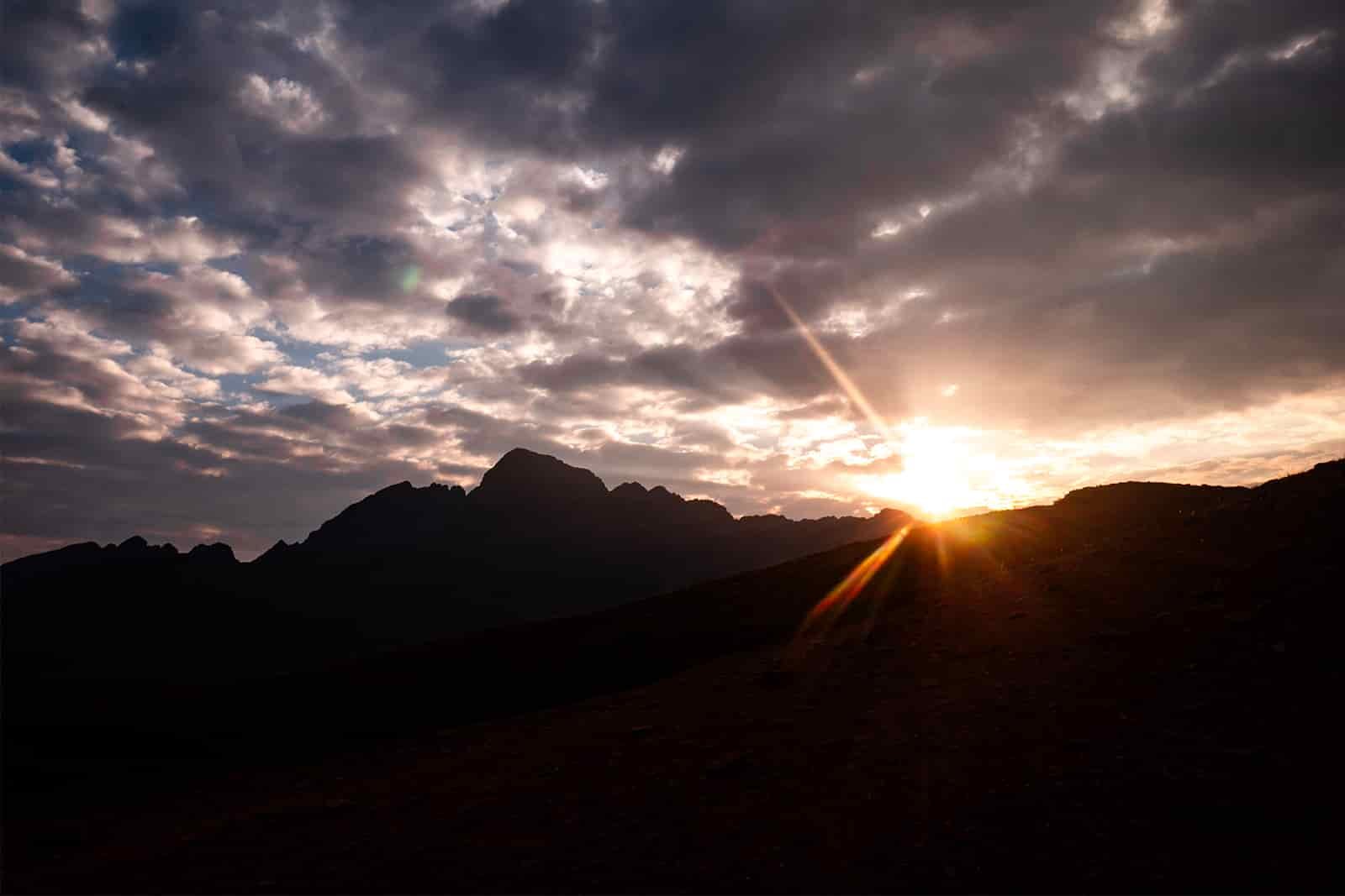 The sun is peaking over the silhouette of ragged mountains. The sky above the sun is cloudy.
