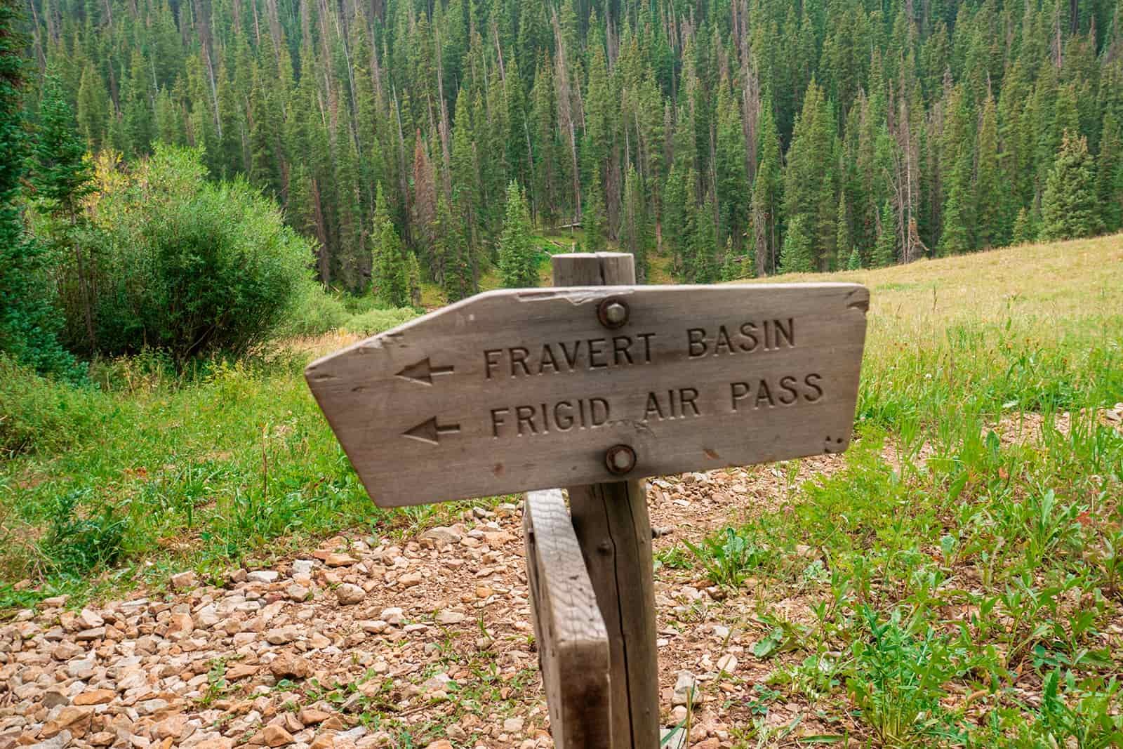 A wooden sign with two arrows pointing to the left read: "Fravert Basin" and "Frigid Air Pass". It is marking a trail junction in the woods.