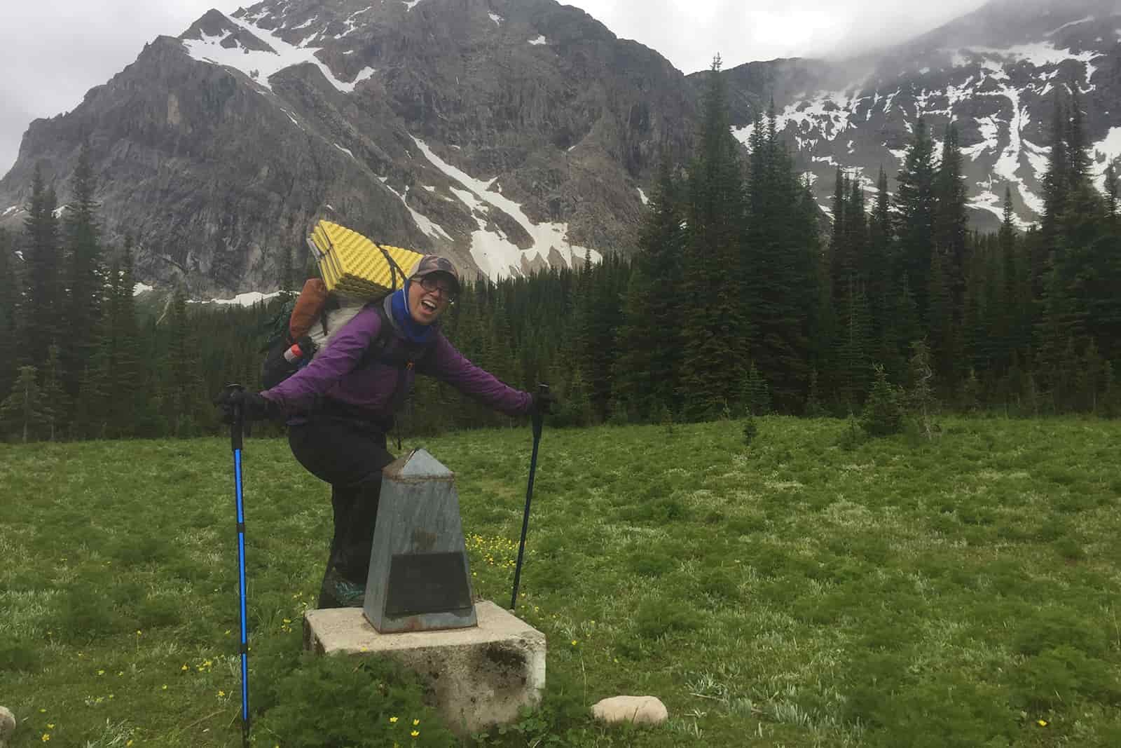 A person wearing a backpack, rain pants, and a rain jacket looking bundled up stands next to a small obelisk. There are pine trees and a mountain in the background and it is a dreary day.