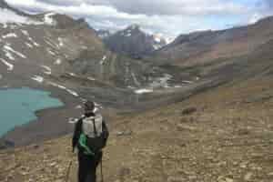 A person wearing a pack is hiking down a scree slope. There is a glacial blue lake in the valley below and mountains in the distance.