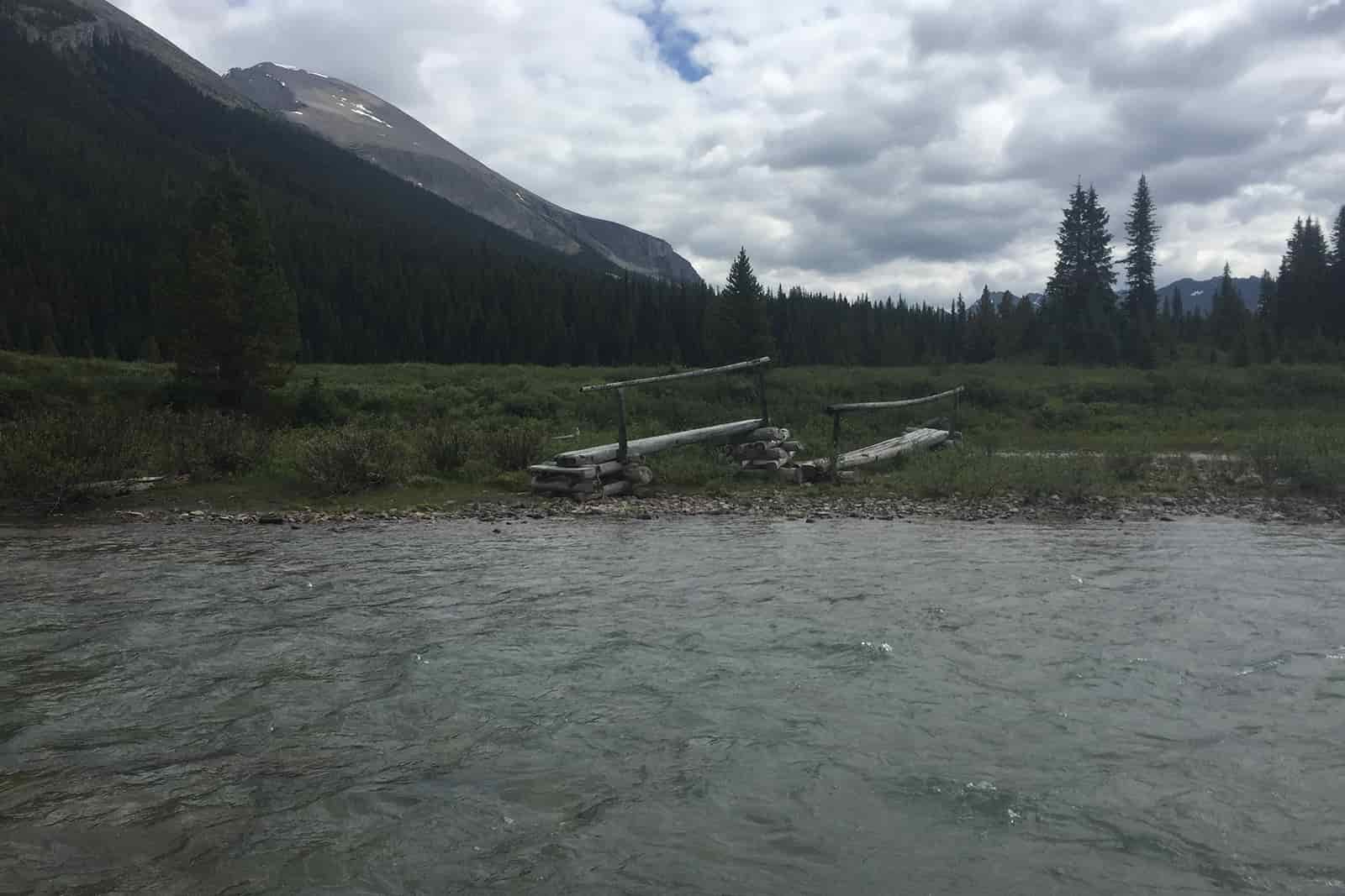 There is a decommissioned bridge on the other side of a fast flowing river. There are trees and mountains in the distance.