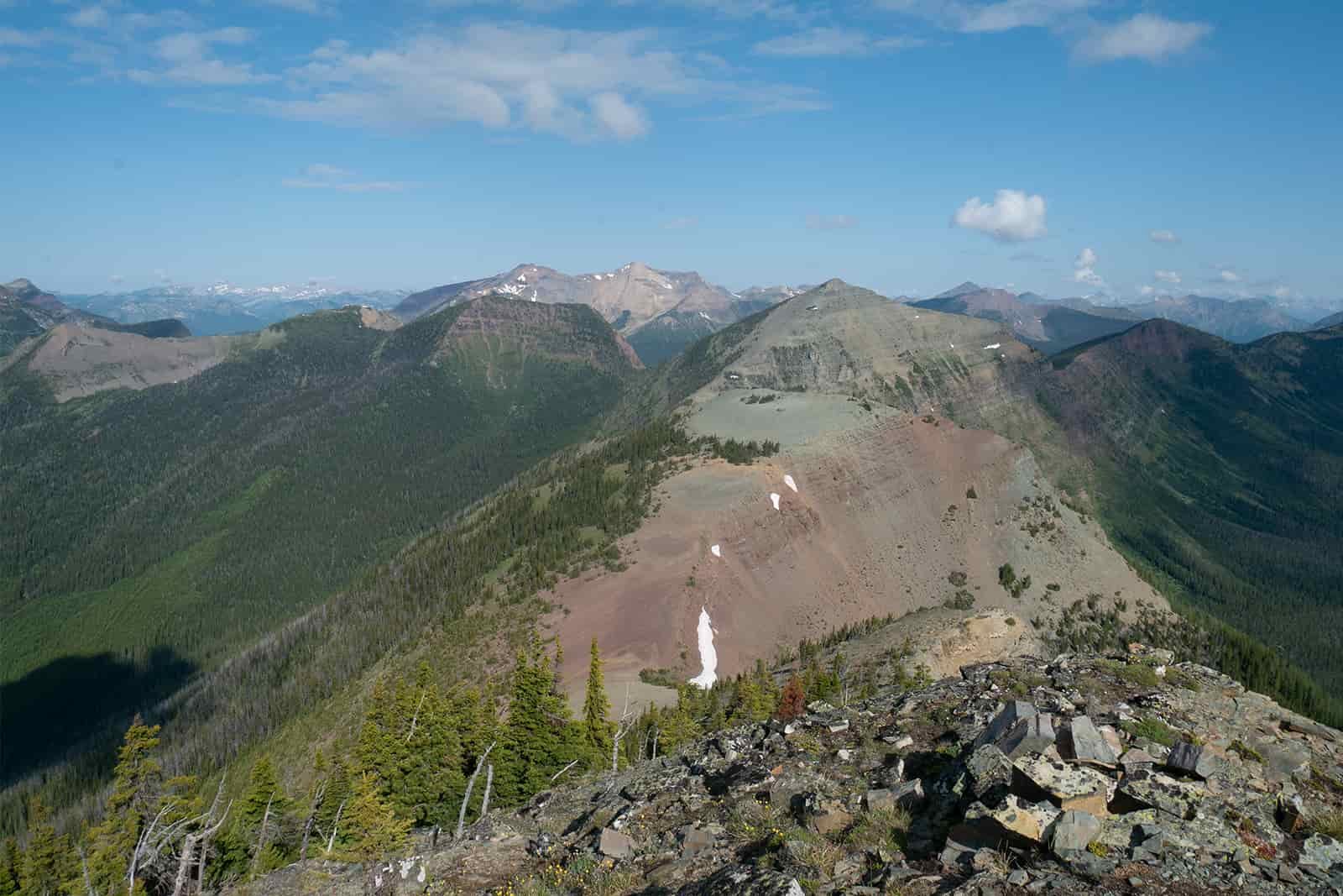 A ridgeline rises and falls steeply between the different peaks. There are other pine covered mountains in the distance.
