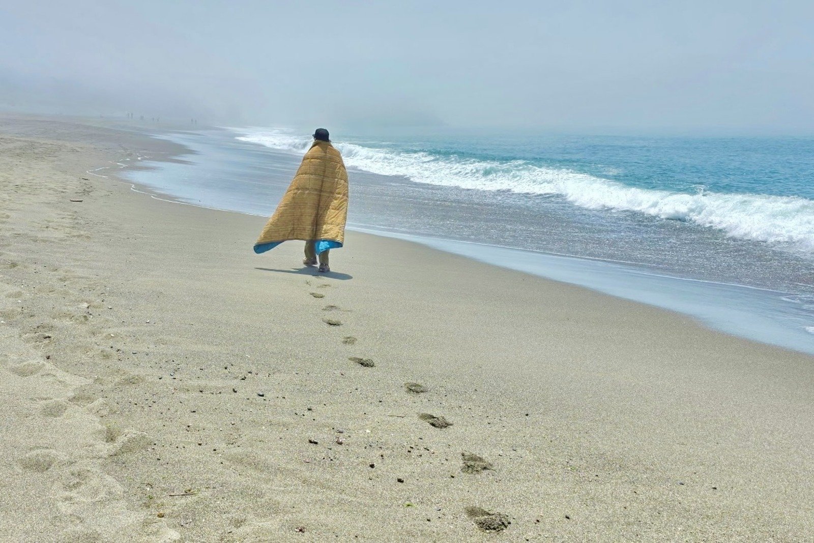 A man walks a long a foggy coastline wearing a blanket.