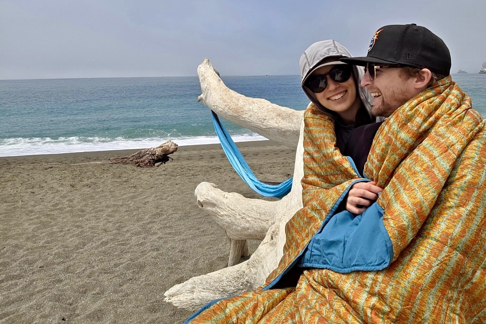 A man and woman sit on a log on the beach, wrapped up in a blanket.