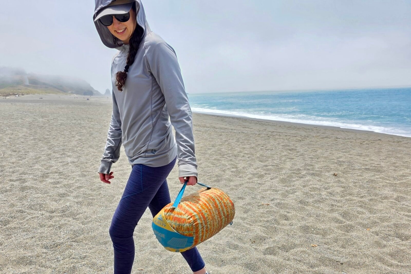 A woman carries her camping blanket across a beach, using the strap on the stuff sack.