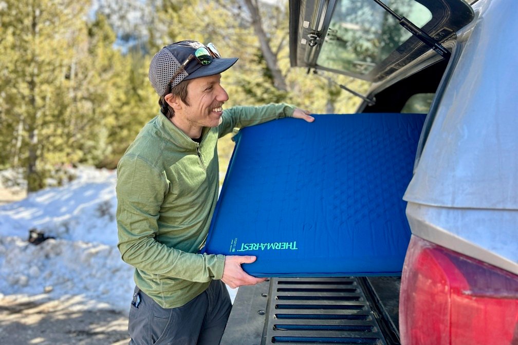 A man smiles as he slides an inflated camping mattress into the back of a pickup truck.