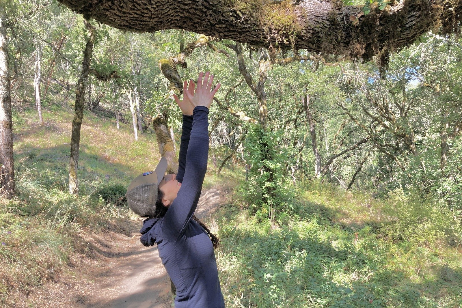 A woman reaches up to a tree branch, showing the side profile of her fleece jacket.