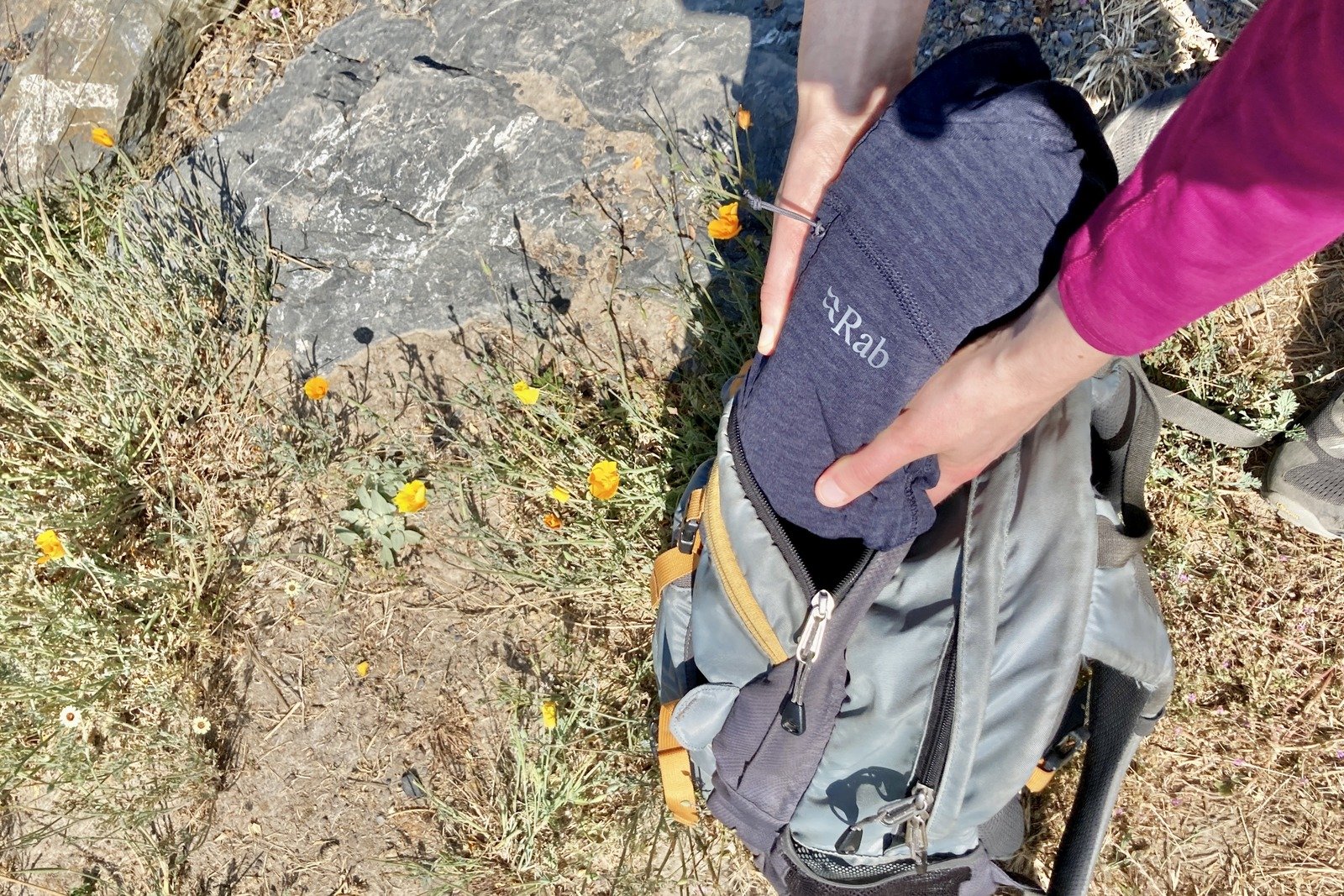 A woman packs up her fleece jacket into her daypack, next to some poppy flowers.