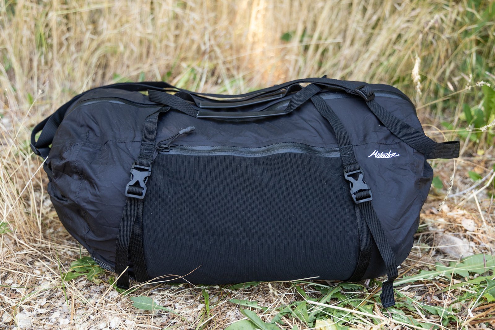 A side view of the Matador Freefly Packable Duffel. You can see two of the side compression straps and buckles, the mesh zipper pocket closed and empty, and the straps resting atop the bag. The matador logo is near the edge.