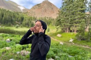 a hiker in a black hooded base layer stands in an alpine environment and raises the hood on his shirt