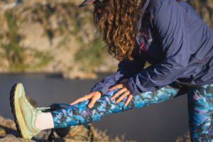 Woman wearing bright blue leggings in a camouflage like pattern on a hike, with one foot up on a rock, stretching her calf and hamstring. She has curly hair and is wearing a purple rain jacket.