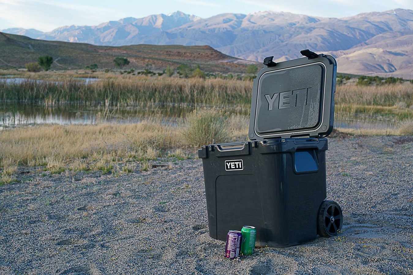 The YETI Roadie 48 cooler sits on sandy ground with its lid open. Two canned drinks sit beside it.