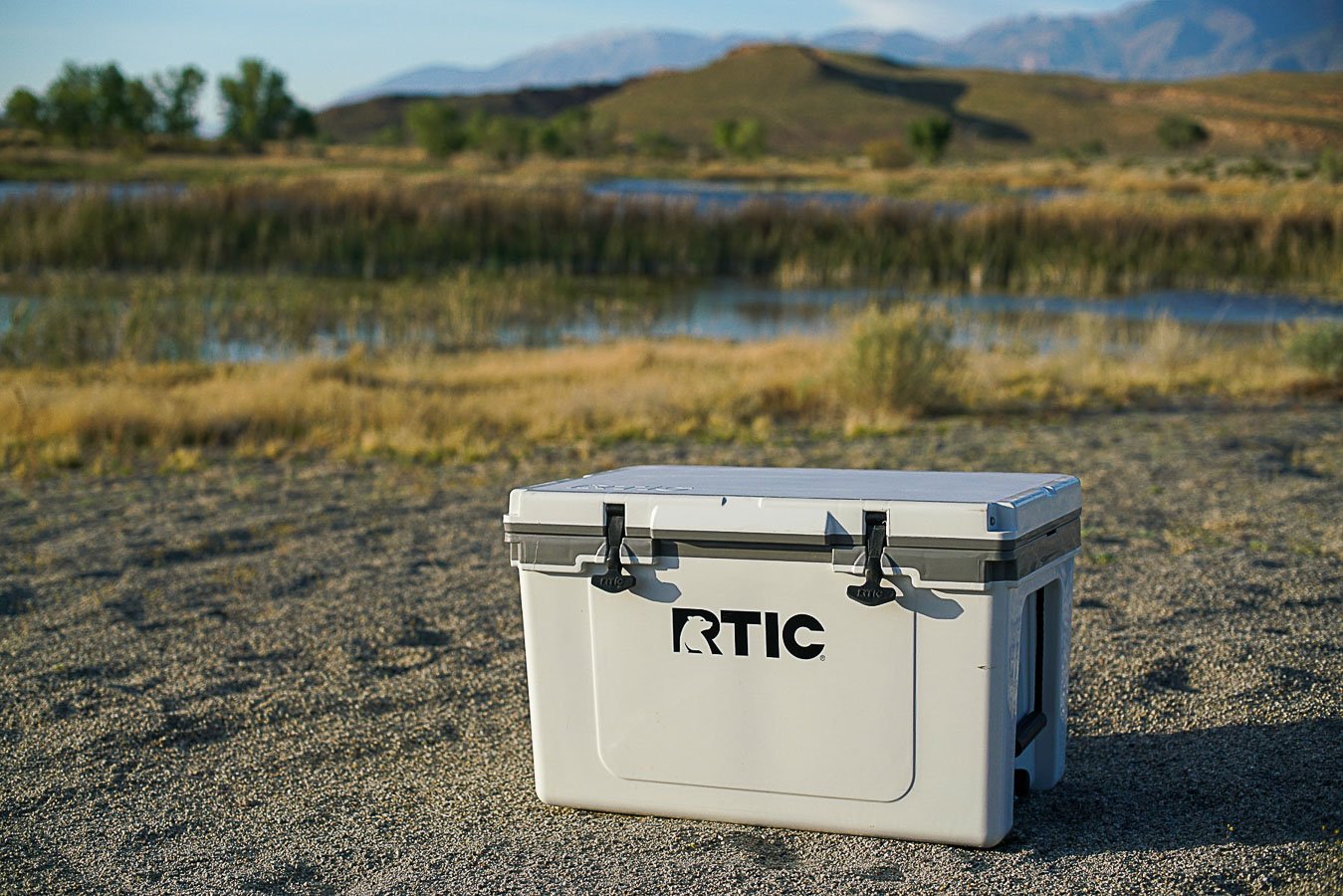 The RTIC Ultralight 52 cooler sits on sandy ground with a body of water and mountains in the background.