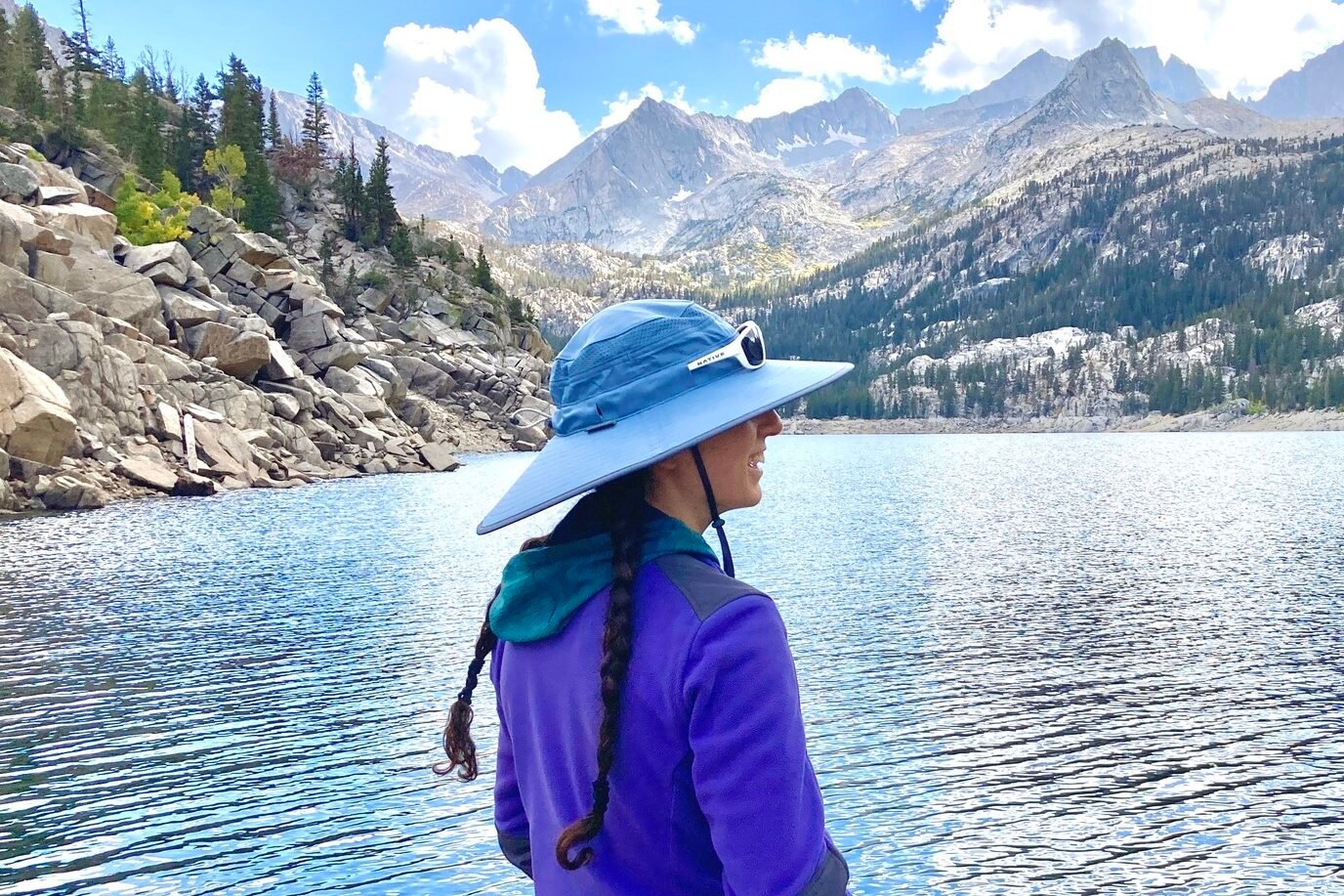 A woman looks out at mountain peaks in the distance in front of a lake, wearing her sun hat.