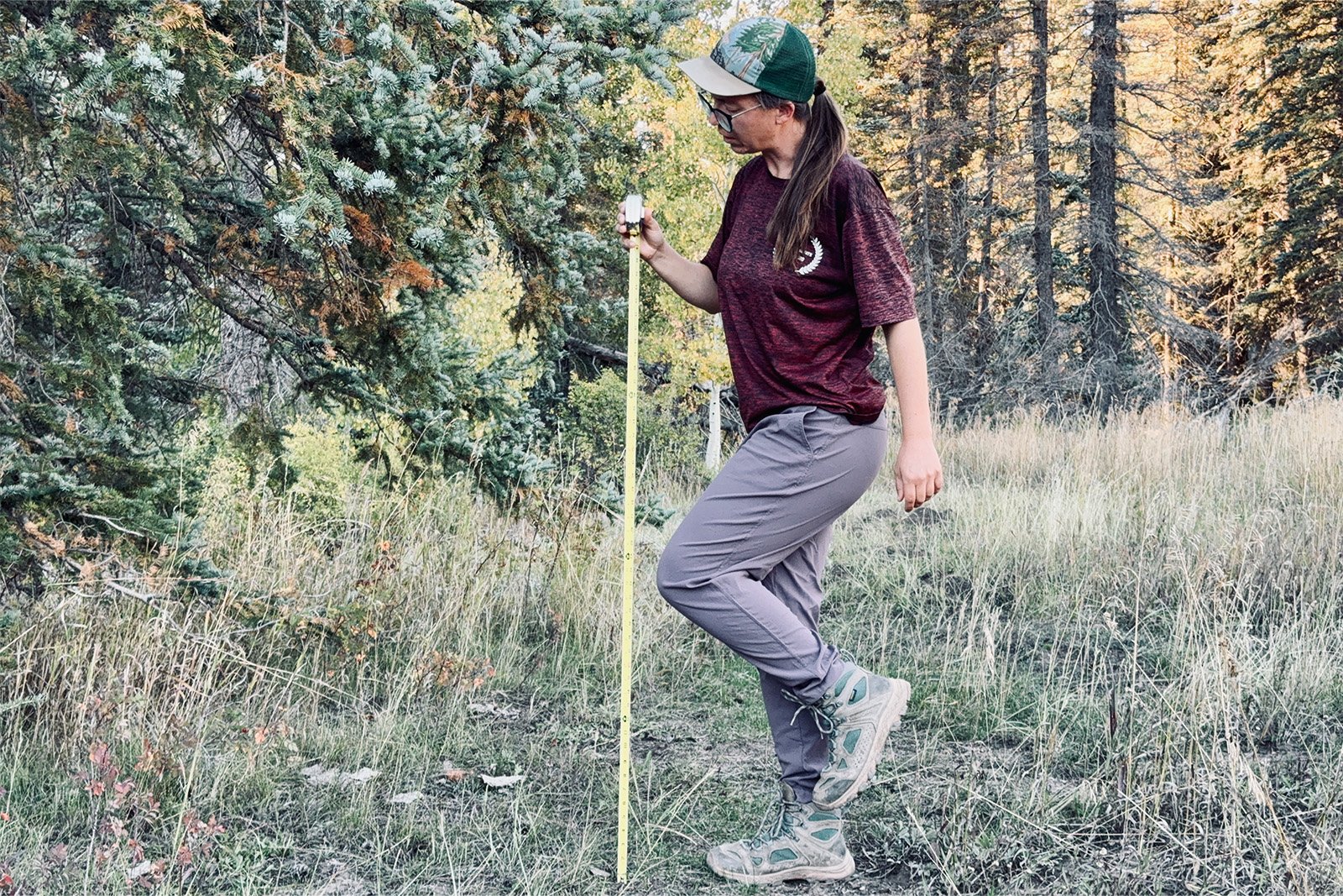 A person is standing on one leg and holding a tape measure up to the other to measure how much mobility the Dynama pants offer. They are standing in the woods.