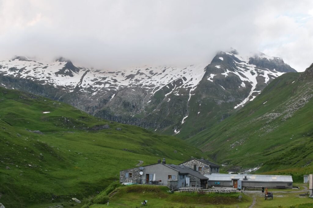 Looking down on stone dorms and metal agricultural buildings in green valley flanked by snowy mountains