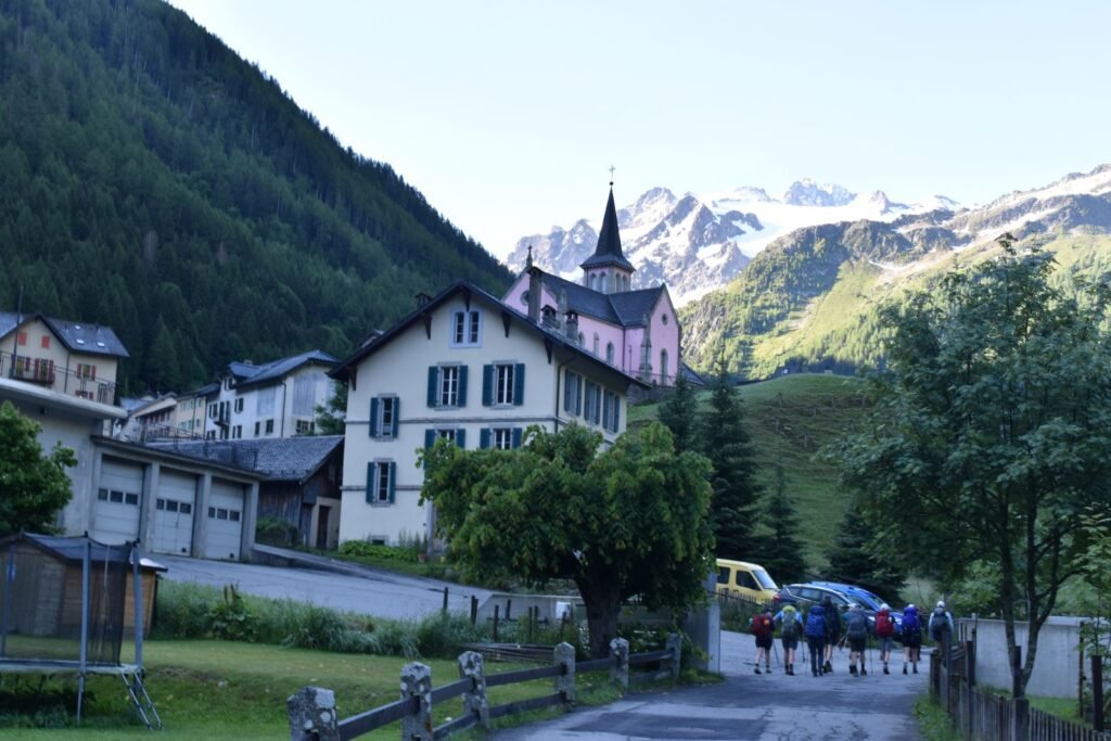 Hikers walk by a pink church and stucco chalets in a Swiss mountain village