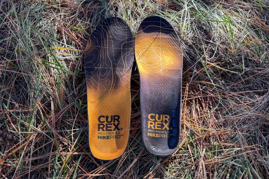 Top view of Currex HikePro insoles lying on dry grass with bold orange and black topo pattern graphics.