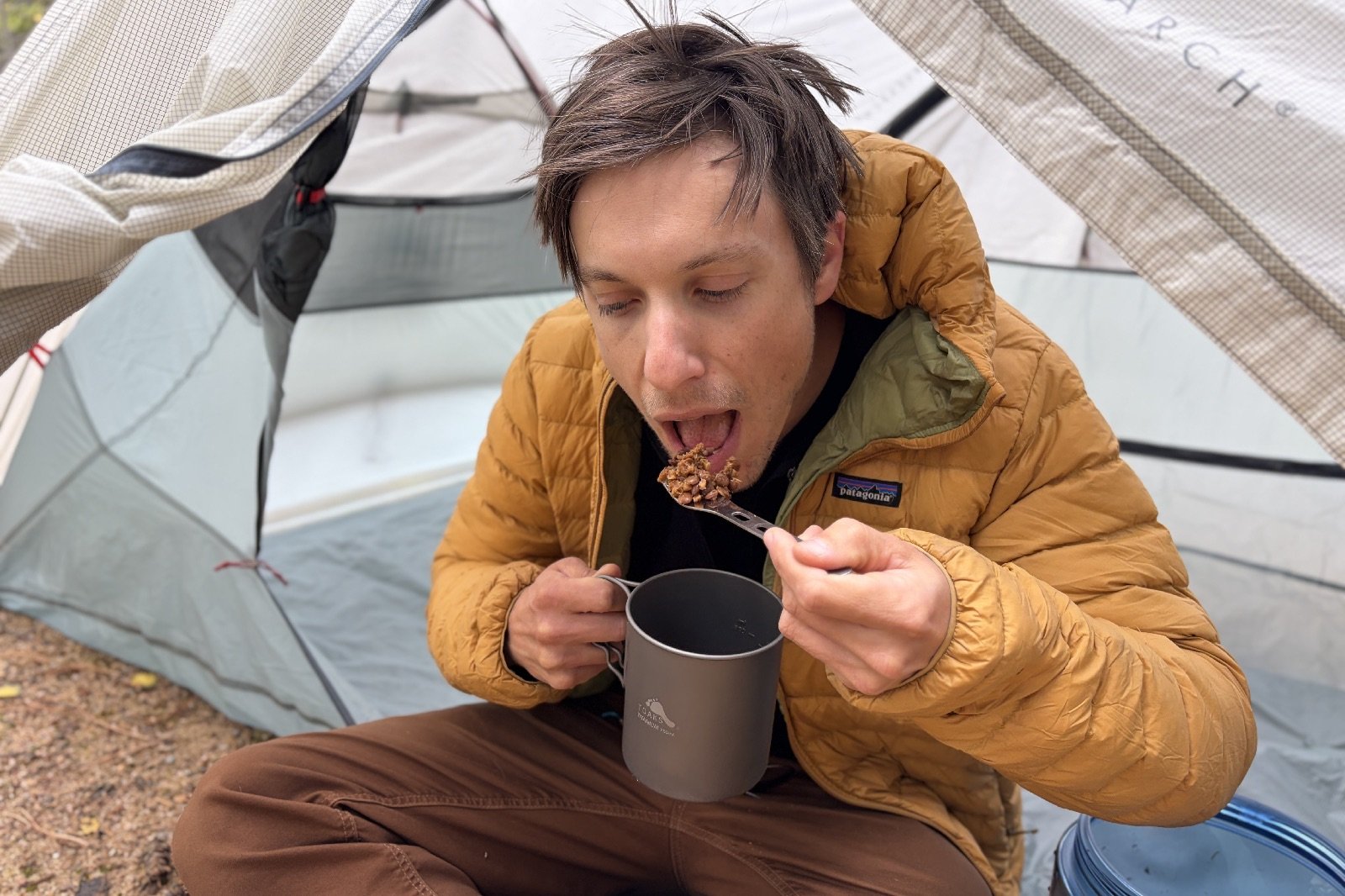 a hiker barbarically shovels hiker food into his mouth with a fork. a tent is in the background.