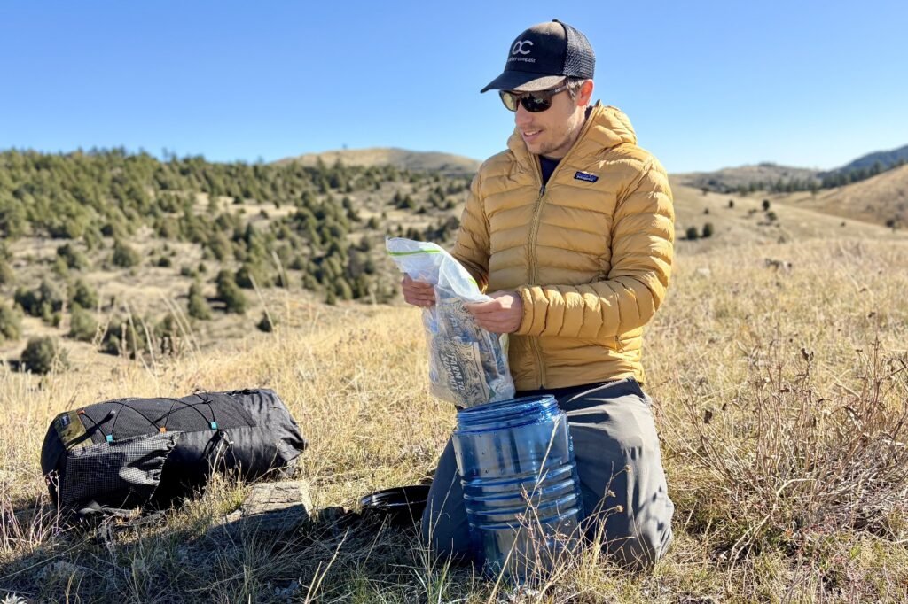 a hiker looks at a clear food bag while kneeling next to a black backpack and blue bear canister