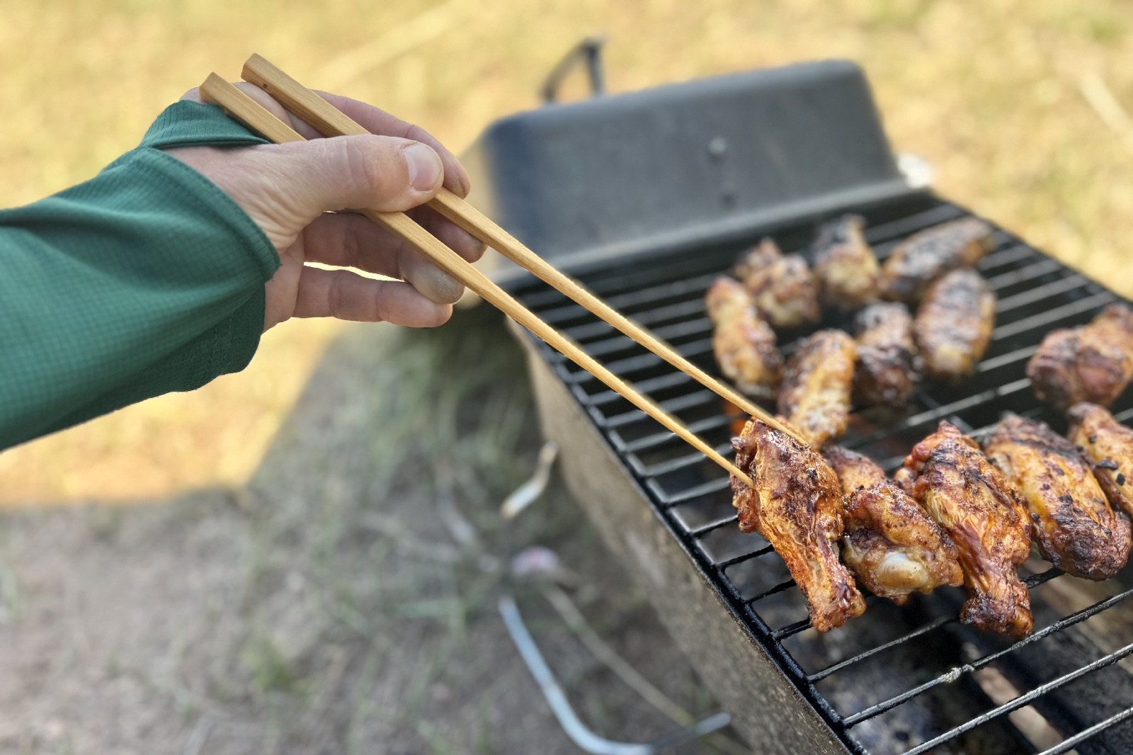 a hand uses chopsticks to pick a chicken wing off of a grill