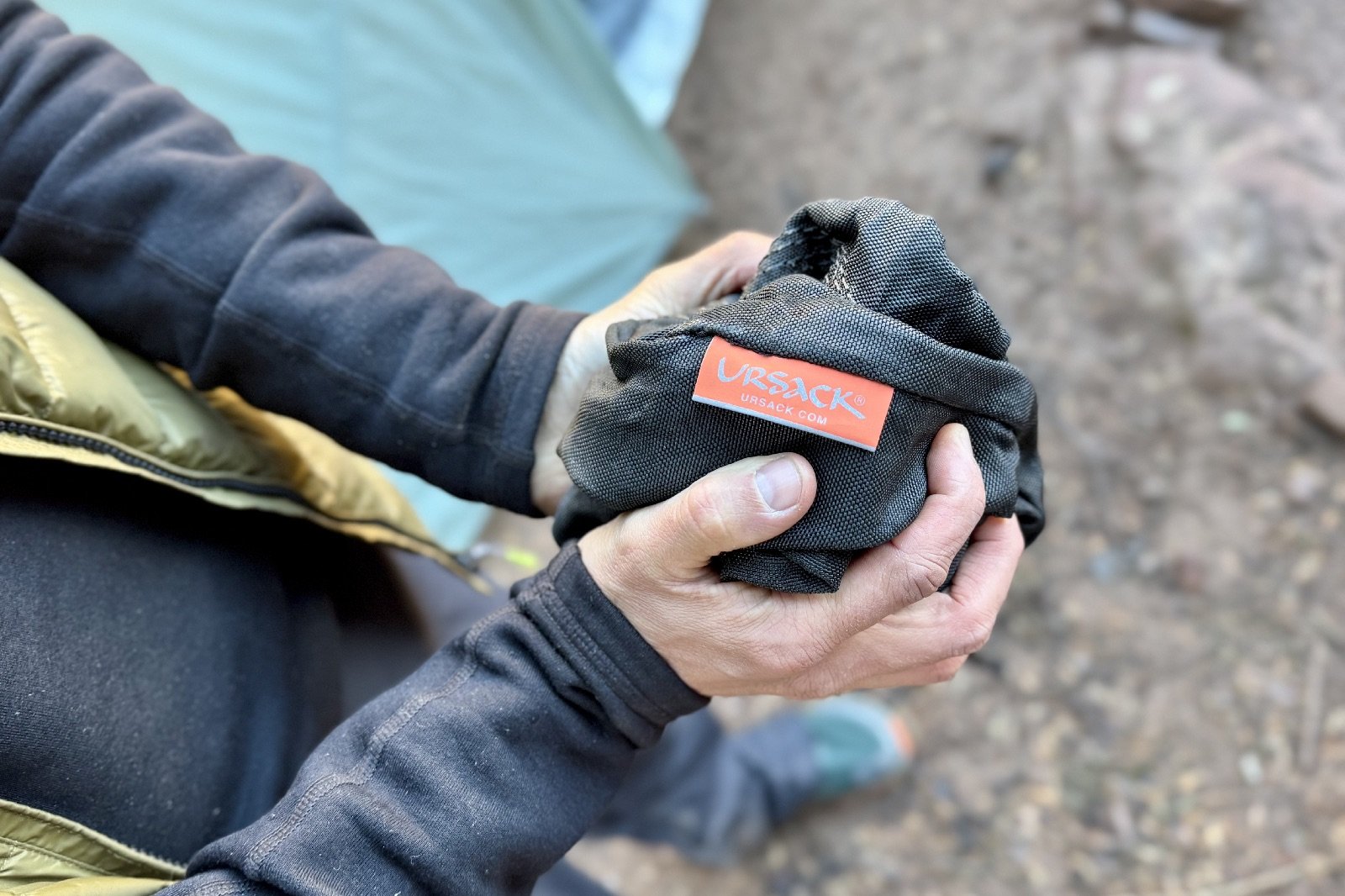 a close up of a hiker's hands balling up a black bag with an ursack tag