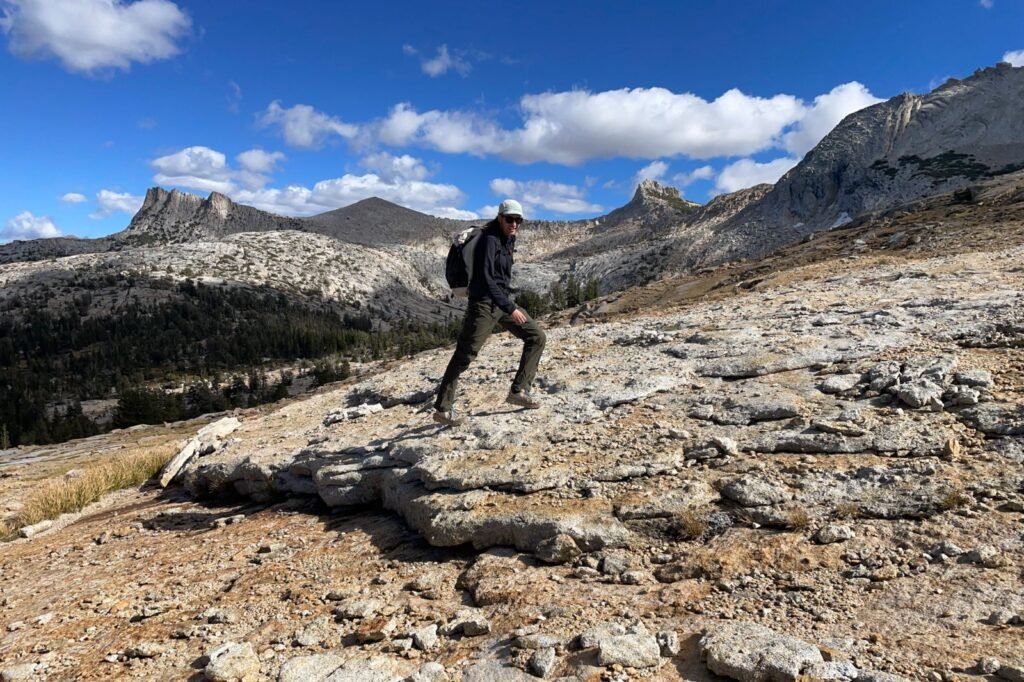 A man walks up a rocky hill in the alpine wearing green hiking pants.