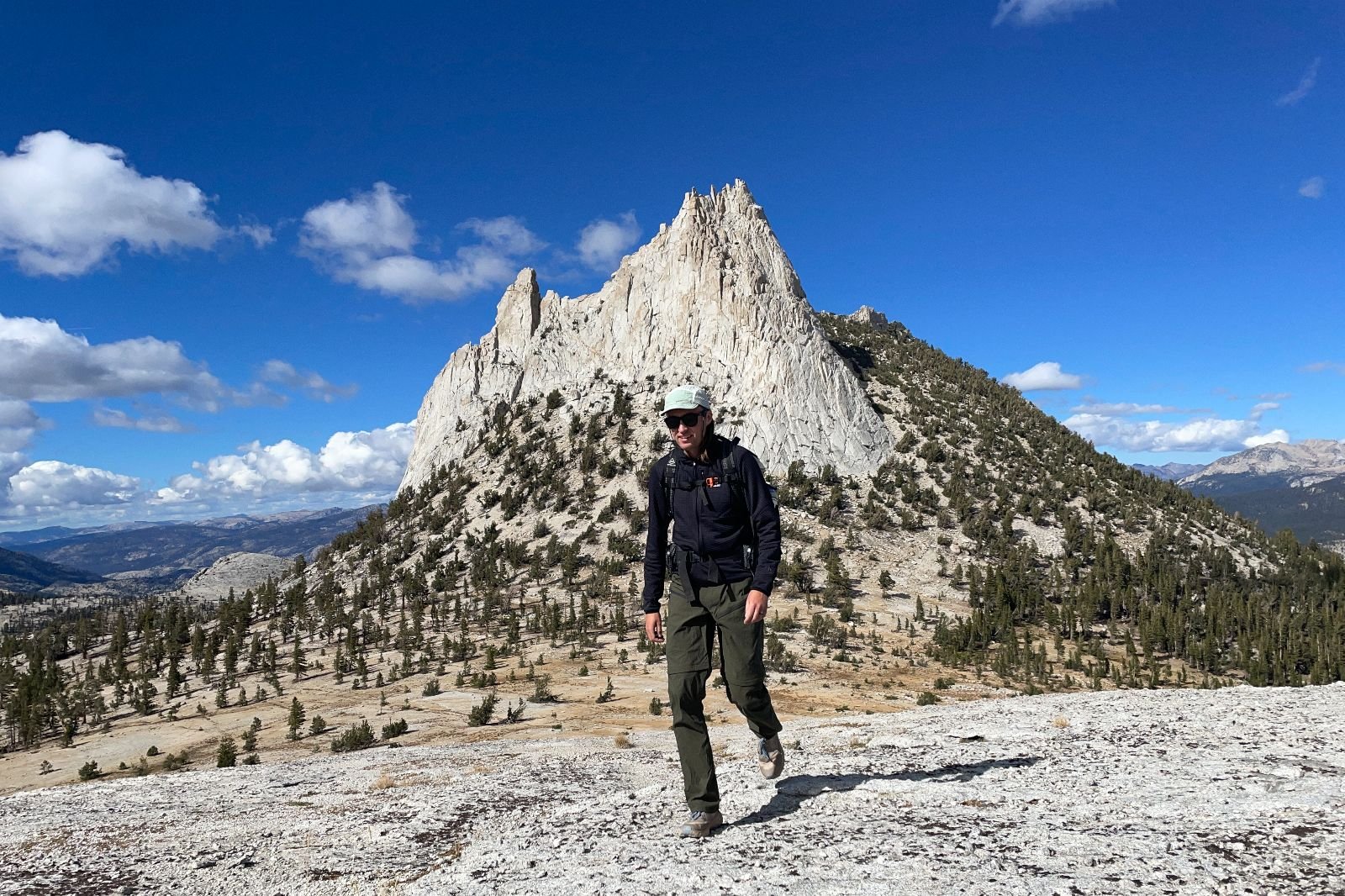 A man hikes in front of a jagged peak while wearing green pants.