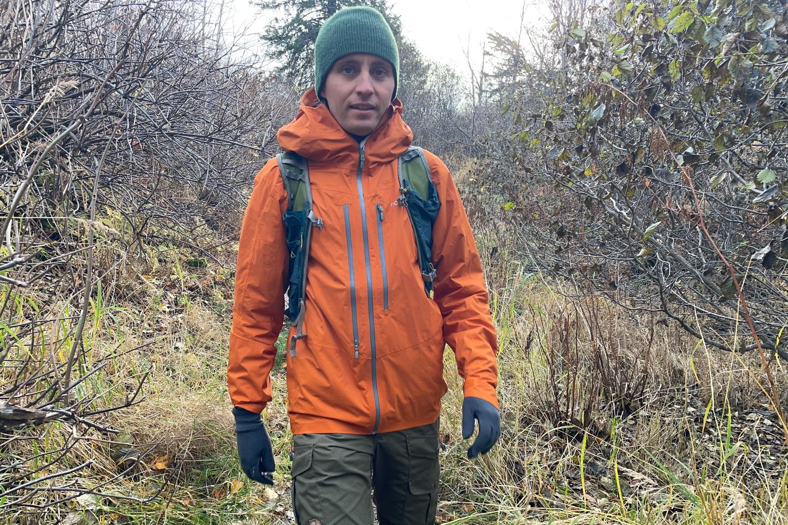 A man hikes while wearing a green backpack and orange rain jacket.