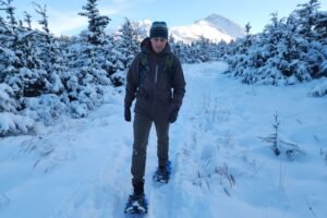 A man snowshoes through a winter landscape with mountains in the background.