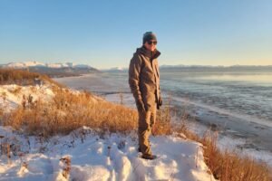 A man stands on a snowy hill overlooking the ocean at sunset.