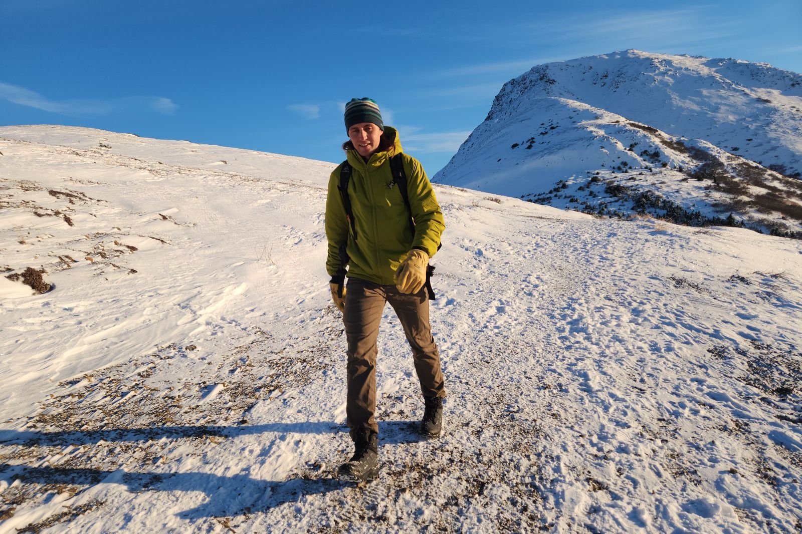 A man walks down a trail in the snowy alpine with a mountain in the background.