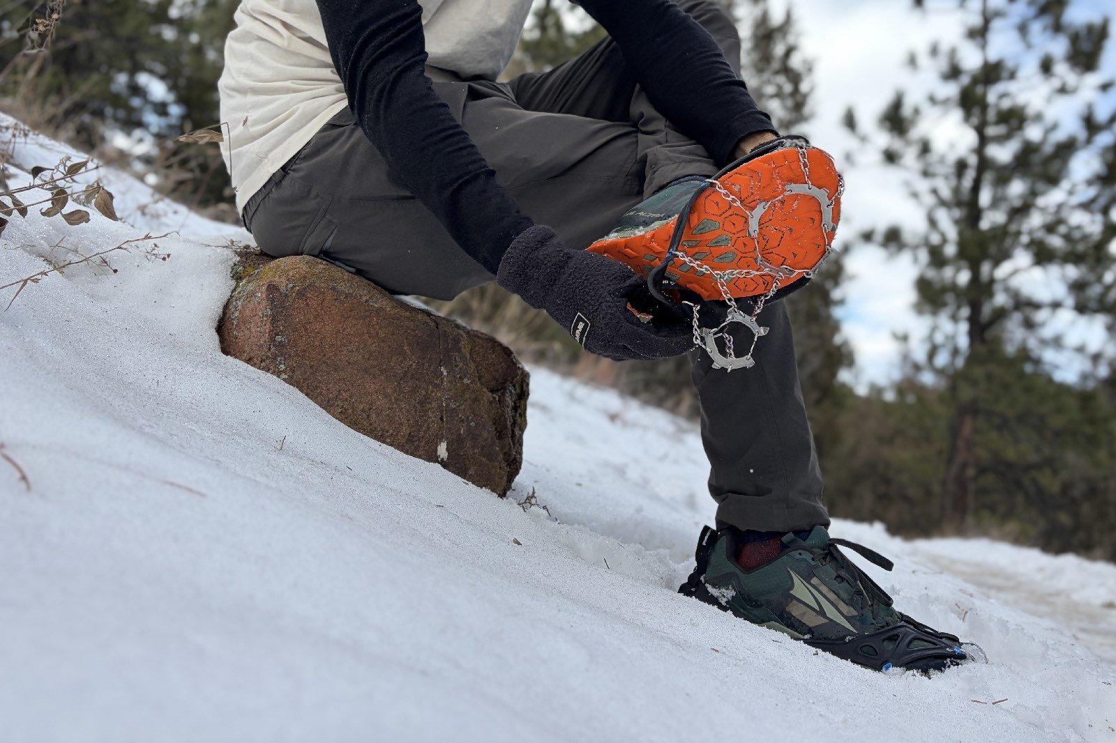 a hiker sits on a rock in the snow and puts on an ice cleat