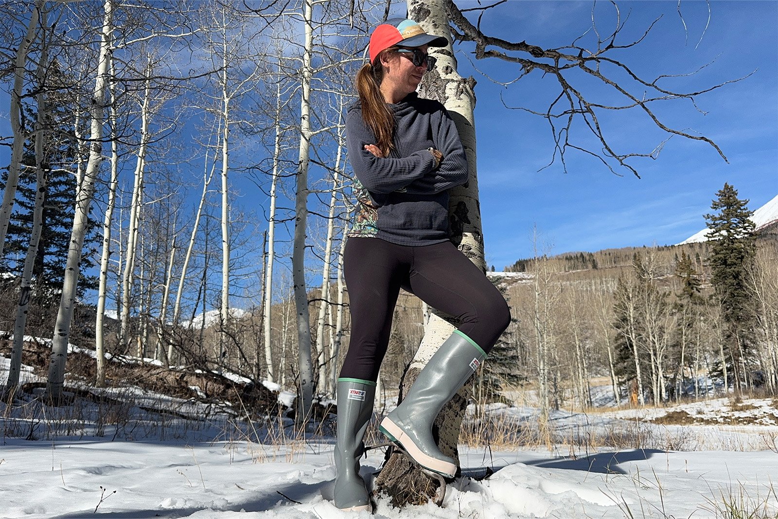 A person wearing Xtratuf boots is leaning against an aspen tree. There is snow on the found and mountains in the background.