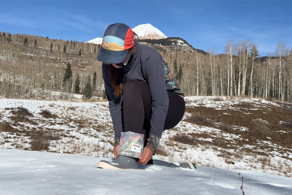 A person is squatting in the snow and rolling down the Xtratuf boots to show the sea themed pattern linin the inside. There is an aspen forest and snow in the background.